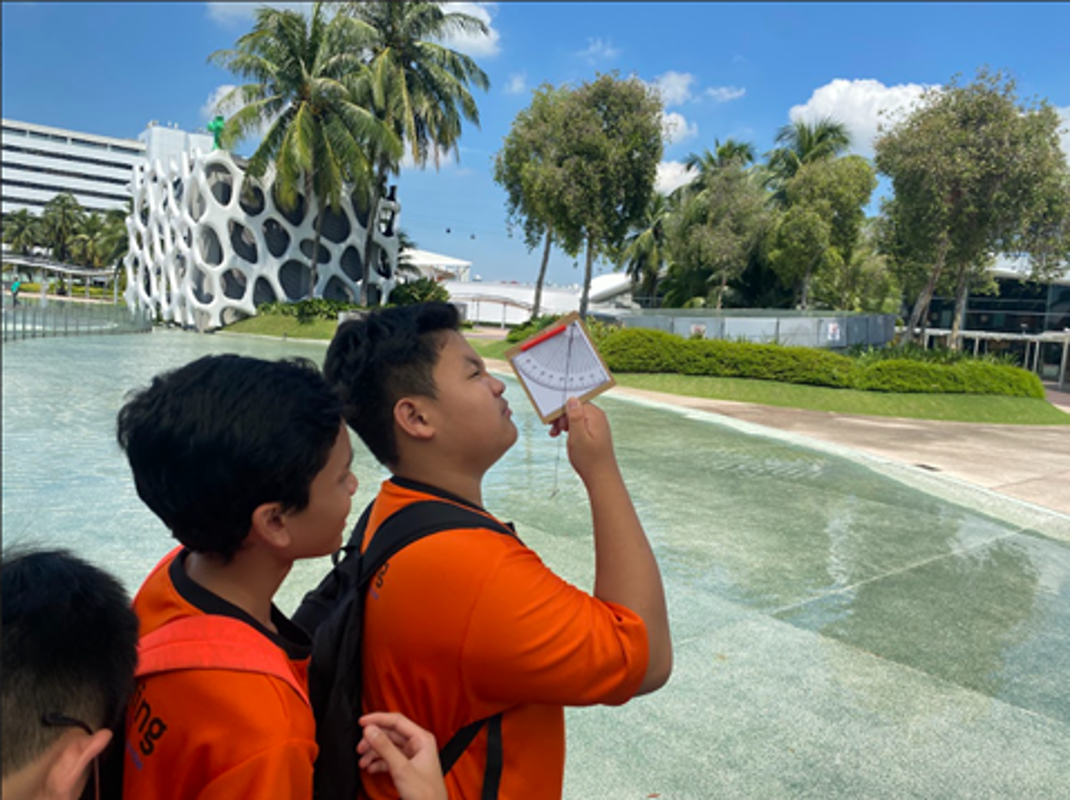 Students outdoors use a handheld clinometer to measure angles while observing their surroundings near a shallow water feature and modern buildings.